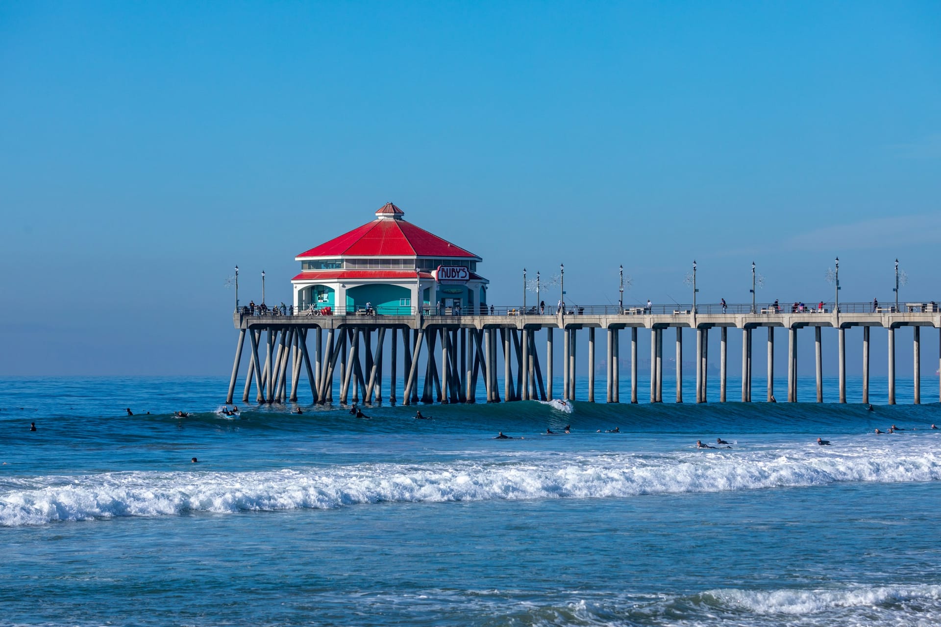 A long concrete pier with a red-roofed octagonal building at its end, above blue ocean waves with small surfers in the water