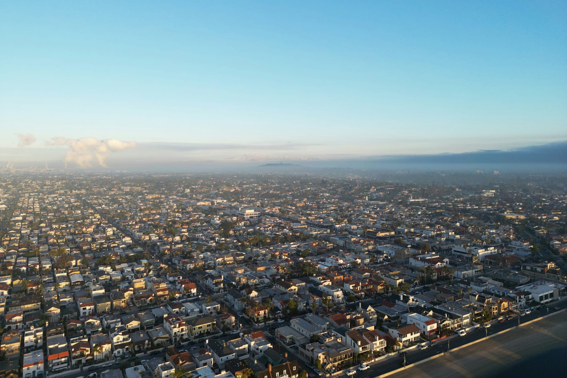 Aerial view over a dense grid of homes and streets beside a beachfront road, under a clear morning sky with a low hill on the hazy horizon