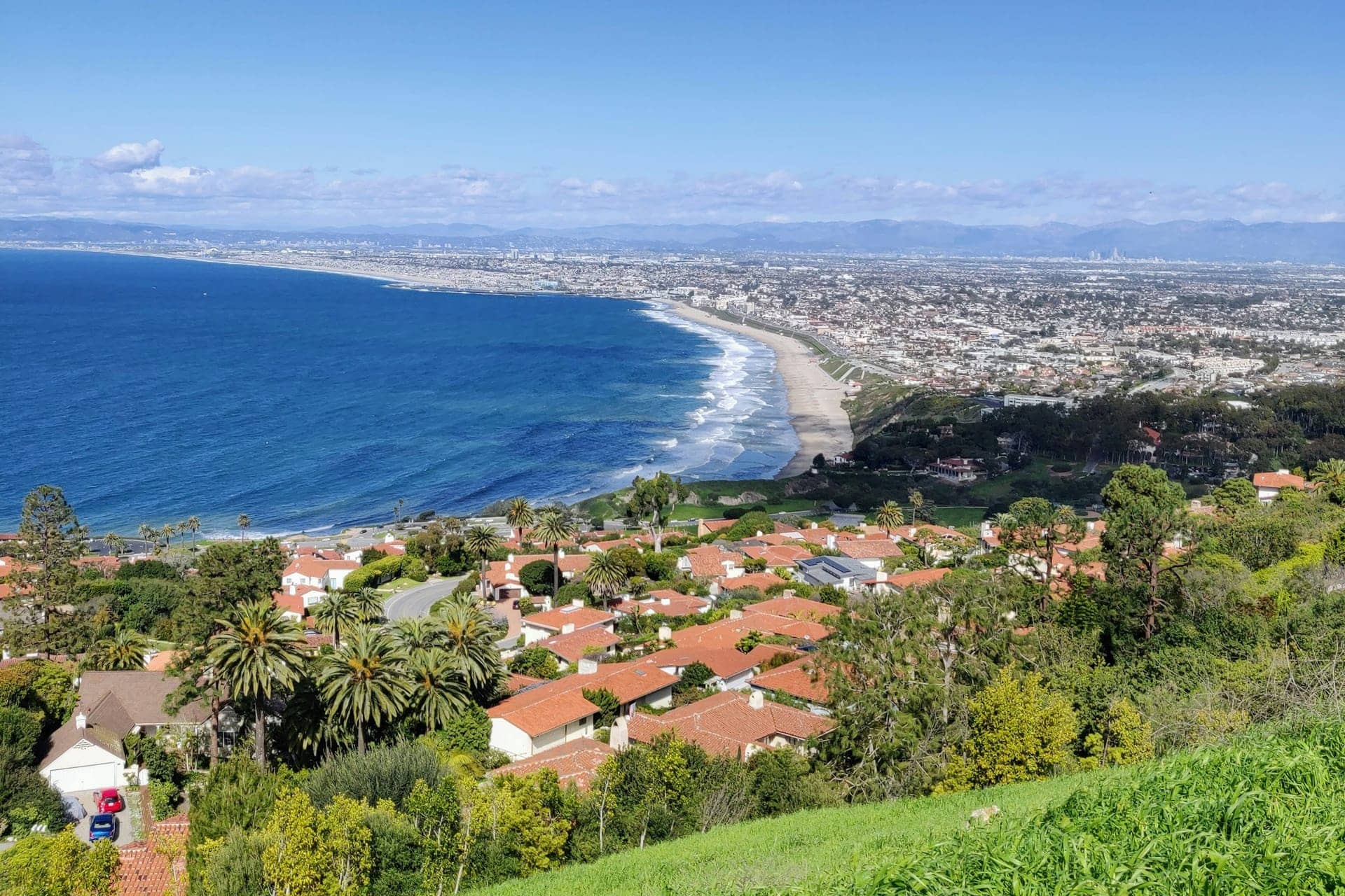 Hillside view over red-tile-roofed homes and palm trees toward a curving sandy beach, blue ocean and a coastal city stretching to distant mountains
